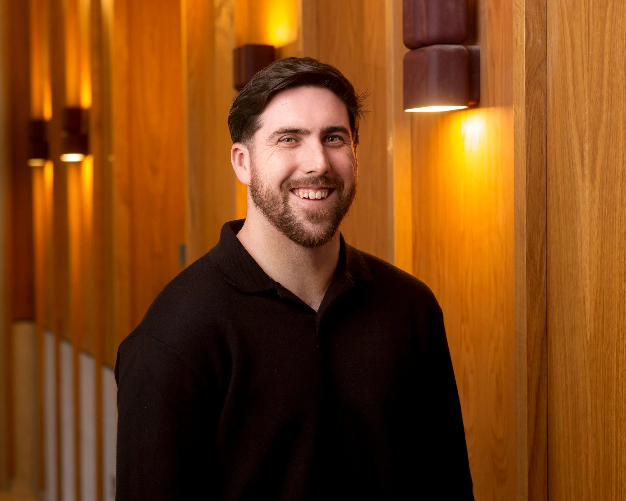 Aaron Duffin Lyons smiling, wearing a black polo, standing in a warmly lit wood panelled corridor.